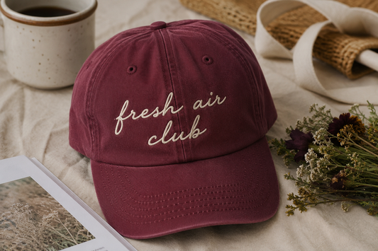 A burgundy dad hat baseball cap with 'fresh air club' embroidered in beige script on the front, styled on a beige fabric surface with a ceramic mug, dried flowers, and a notebook.