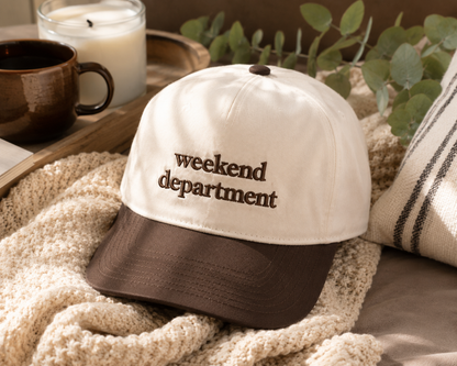 A two-tone baseball cap with an off-white crown and a brown brim, embroidered with the words 'weekend department' in brown. The cap is styled on a cozy beige knit blanket with a brown mug, a candle, and green leaves in the background.