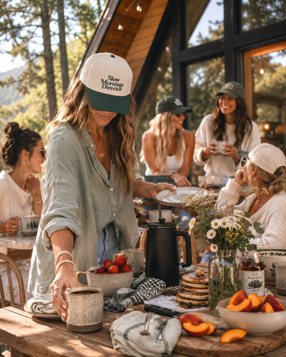 A lifestyle scene at a rustic A-frame cabin where a woman in a light blue shirt and a white cap with green trim and the text 'Slow Mornings Director' is serving coffee. Other women are gathered around the table with fruit, flowers, and mugs in a warm, sunny outdoor setting.