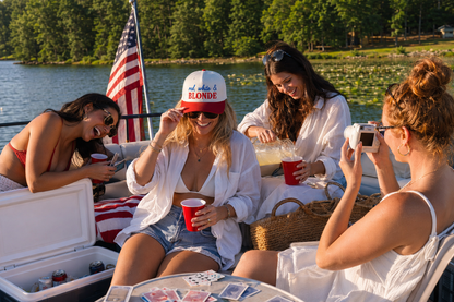 Four women on a boat by a lake wearing matching white linen shirts, holding red cups, and laughing together. One woman wears a beige and red baseball cap with 'red white and BLONDE' printed on it.