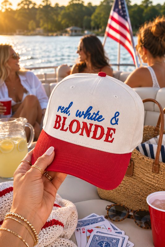 A hand holds a red and natural baseball cap in the foreground with the text 'red, white & BLONDE' embroidered on the front. The background shows a sunny outdoor boating scene with people on a boat near the water.