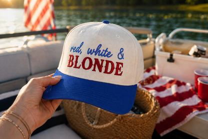 A hand holds a natural and blue trucker-style cap with the text 'red, white & BLONDE' embroidered on the front, set against a sunny boating scene on the water.
