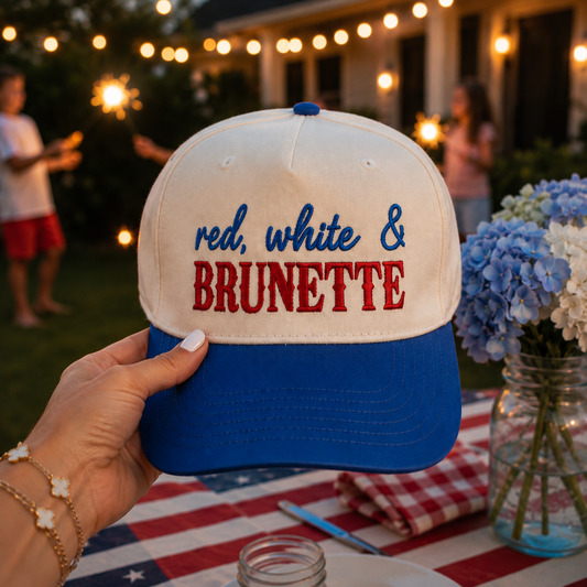 A hand holds a natural and blue baseball cap with the text 'red, white & BRUNETTE' embroidered on the front, set against a backyard party background with people holding sparklers and a table decorated with an American flag napkin and blue hydrangeas.