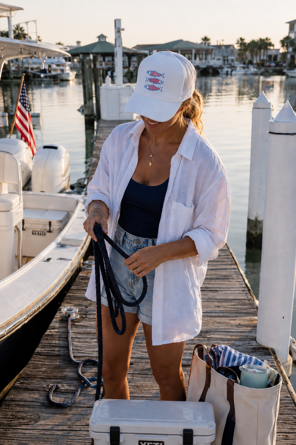 A woman stands on a wooden dock by the water wearing a white baseball cap with a red and blue sardine fish graphic, a white button-down shirt, a black tank top, and denim shorts. She is holding a black rope near a white cooler and a tote bag, with boats and a marina visible in the background.