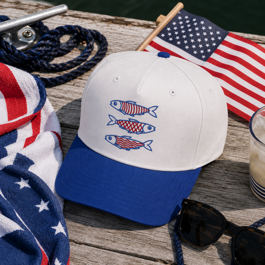 A white and royal blue baseball cap with three embroidered red, white, and blue fish on the front, styled on a wooden dock with an American flag, sunglasses, a towel, and rope nearby.