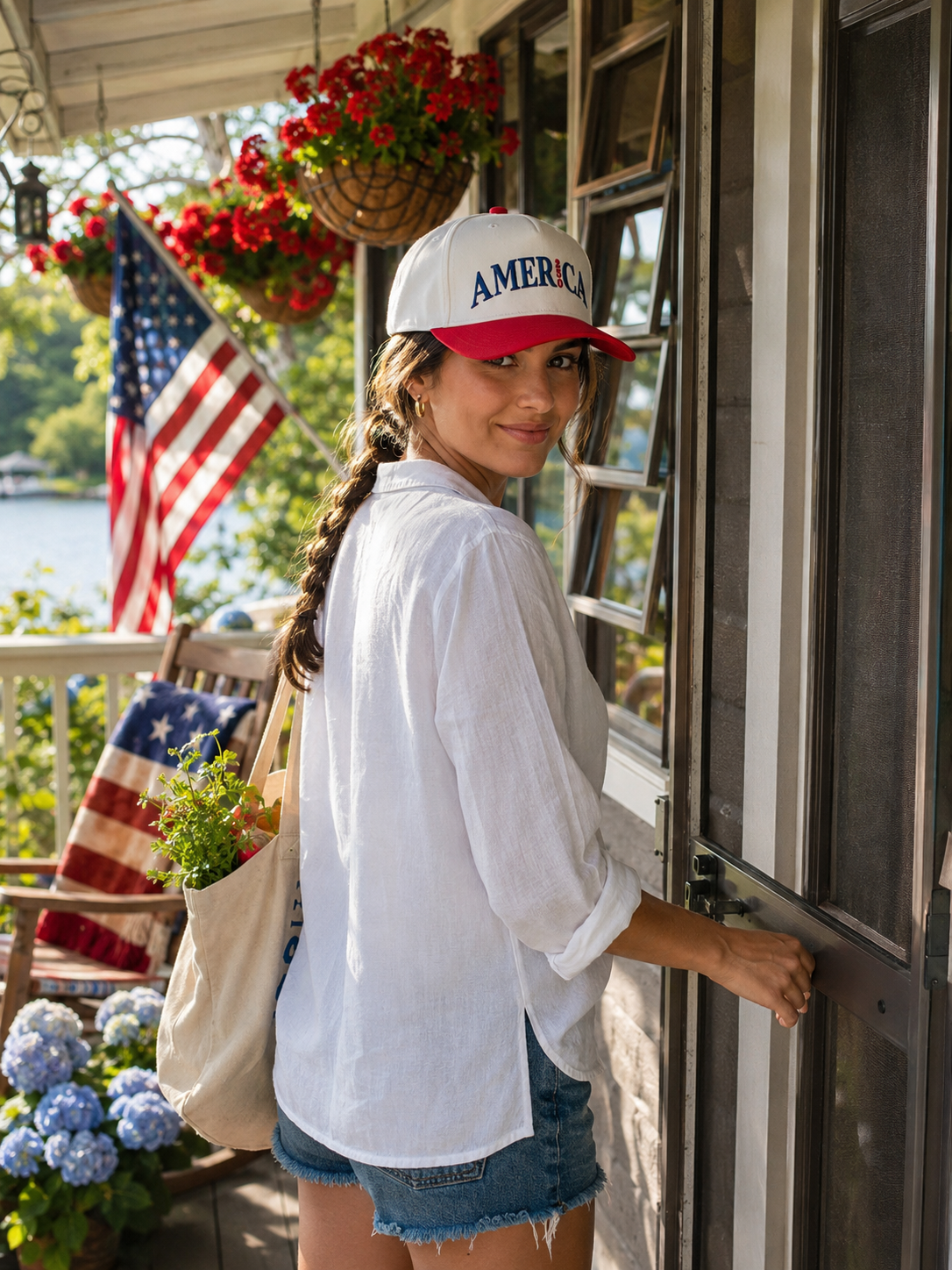 A woman wearing a white and red baseball cap with 'AMERICA 250' printed on the front, a white button-up shirt, and denim shorts stands on a porch by a window.