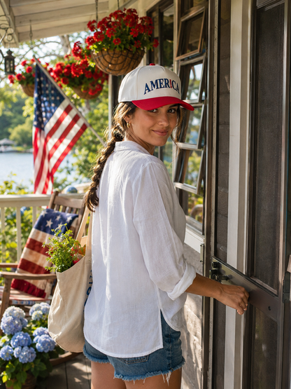 A woman wearing a white and red baseball cap with 'AMERICA 250' printed on the front, a white button-up shirt, and denim shorts stands on a porch by a window.