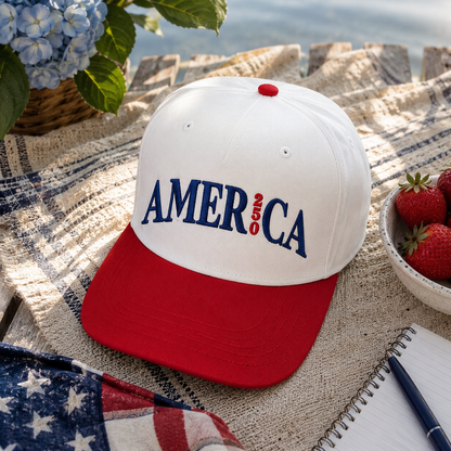A red and white baseball cap with 'AMERICA 250' embroidered on the front, styled on a picnic blanket with strawberries, flowers, and an American flag in the background.