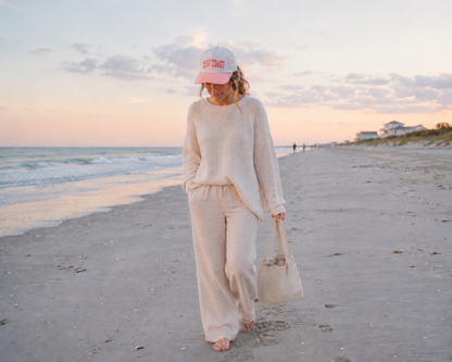 A woman walks barefoot on a sandy beach at sunset wearing a cream knit lounge set consisting of a long-sleeve top and matching wide-leg pants, a white baseball cap with red text, and carrying a beige tote bag.