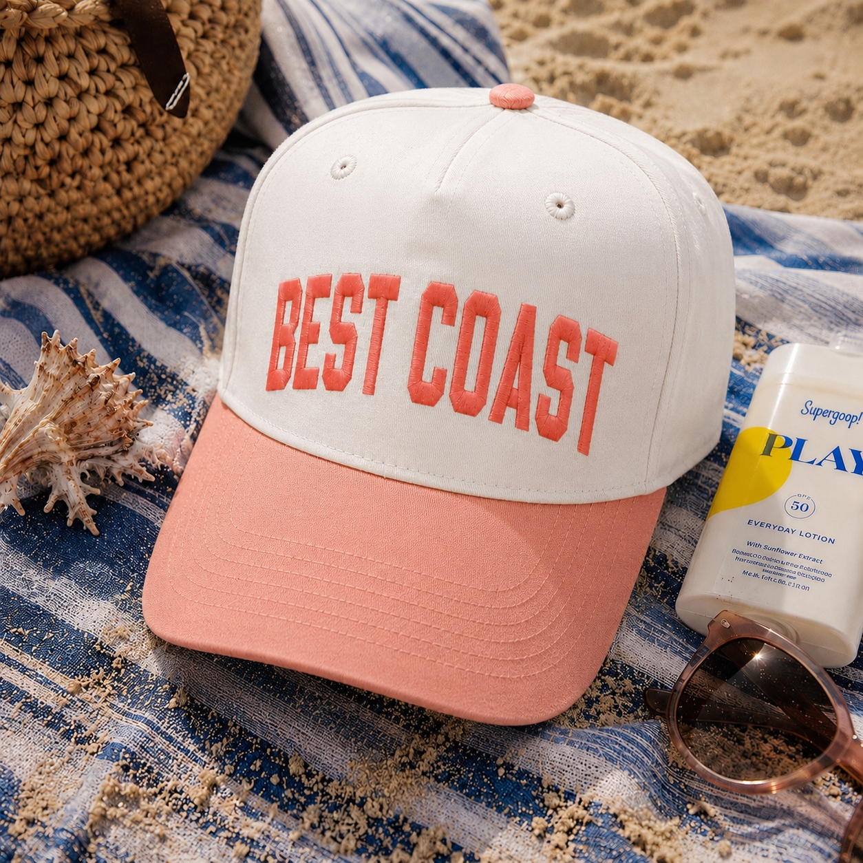 A natural baseball cap with a coral-pink brim and 'BEST COAST' embroidered on the front, styled on a blue-and-white striped beach towel with sand, sunglasses, sunscreen, and a woven hat nearby.
