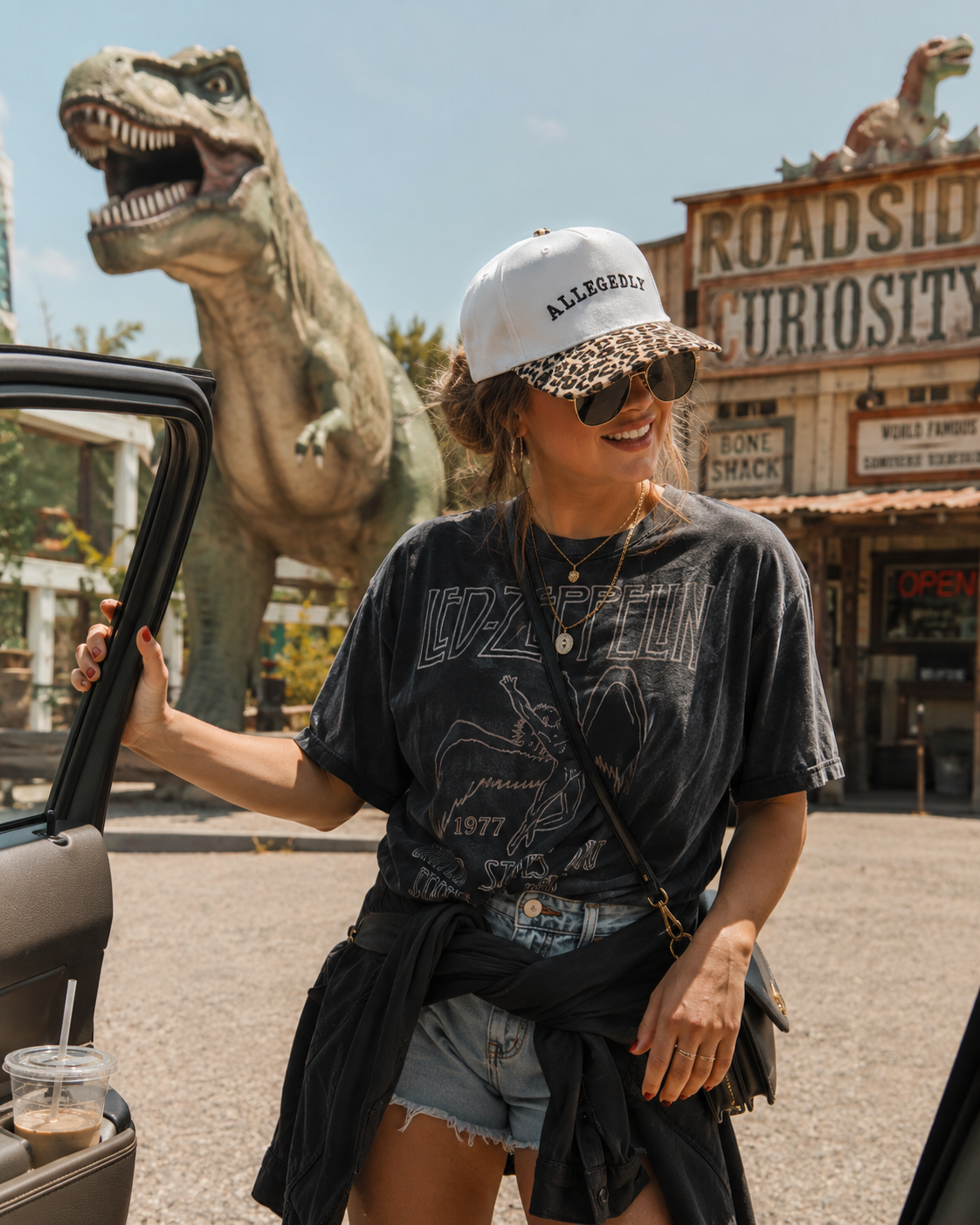 A woman wearing a natural cheetah print baseball cap that has ‘allegedly’ embroidered on the front,  sunglasses, a black graphic t-shirt, and denim shorts stands by an open car door in front of a dinosaur statue and a vintage-style diner sign.