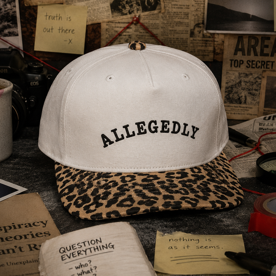 A beige baseball cap with 'ALLEGEDLY' embroidered in black on the front, featuring a leopard-print visor. The cap is styled on a dark tabletop surrounded by vintage-themed props including a coffee mug, sticky notes, and newspaper clippings.