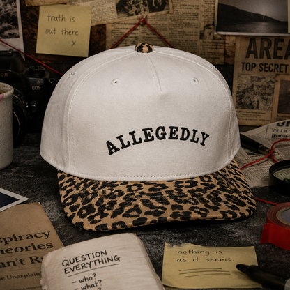 A beige baseball cap with 'ALLEGEDLY' embroidered in black on the front, featuring a leopard-print visor. The cap is styled on a dark tabletop surrounded by vintage-themed props including a coffee mug, sticky notes, and newspaper clippings.