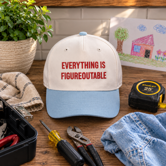 A baseball cap with a cream crown and light blue visor, featuring the red embroidered text 'EVERYTHING IS FIGUREOUTABLE' on the front. The cap is styled on a wooden workbench surrounded by tools, including a tape measure, screwdriver, pliers, a cloth, and a blue denim jacket, with a potted plant and a child's drawing in the background.