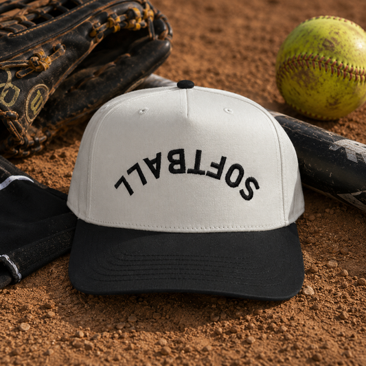 A baseball cap with a white crown and black brim, featuring the word 'SOFTBALL' embroidered in black across the front. The cap is styled on a dirt surface with a softball, baseball glove, bat, and glove in the background.