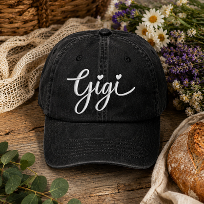 A black washed baseball cap with the name 'Gigi' in white cursive script on the front, styled on a rustic wooden surface with dried flowers, greenery, lace, and bread.