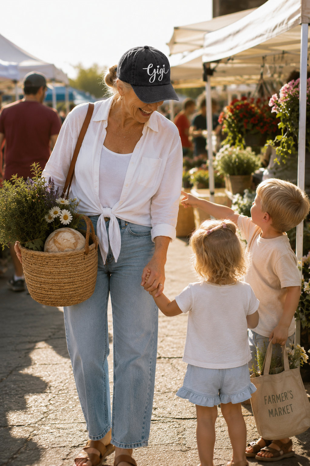 A woman and two children walking at an outdoor farmers market. The woman wears a black baseball cap with the white script logo 'Gigi’ a white shirt tied at the waist, and light blue jeans, while carrying a woven basket.