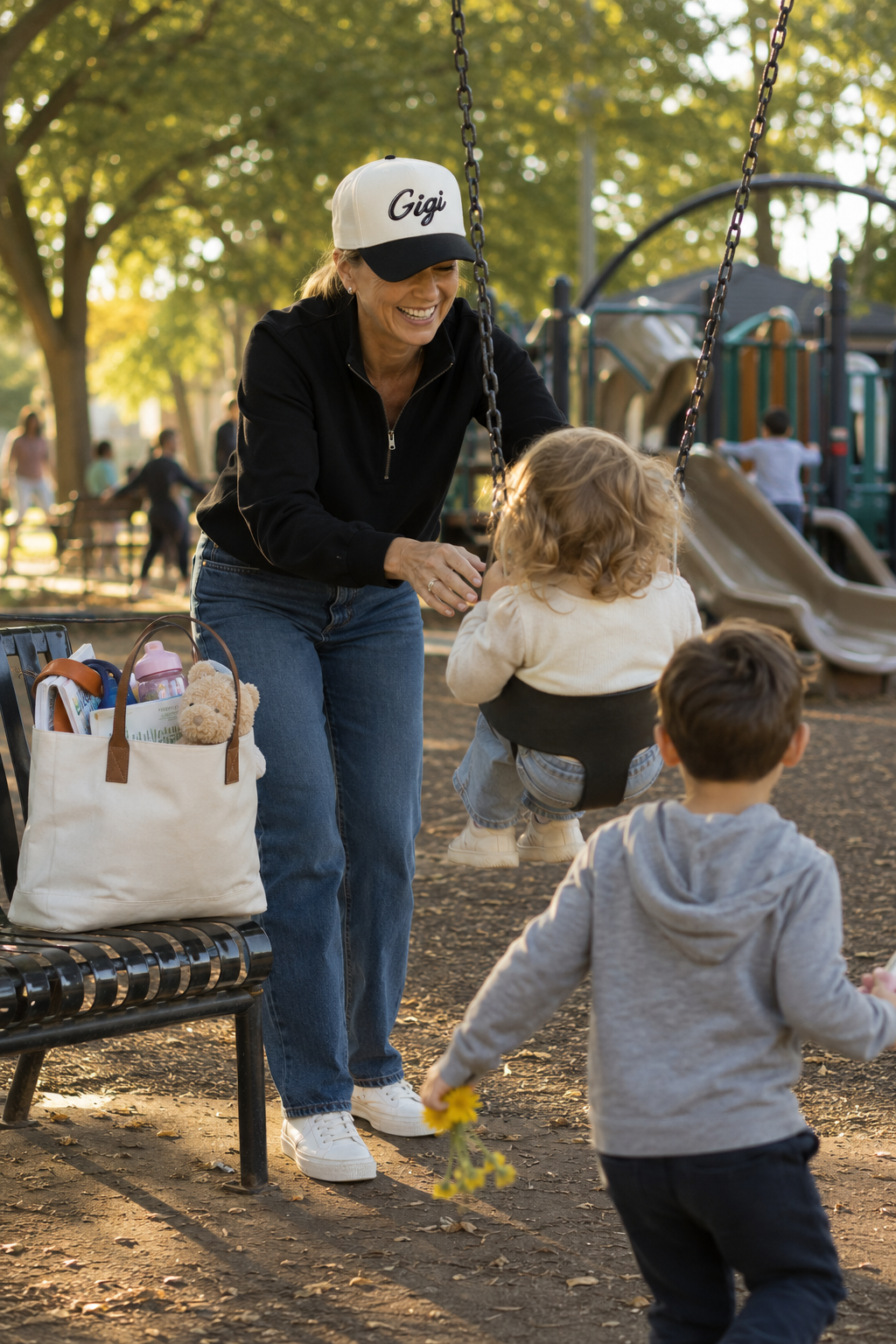 A woman wearing a natural and black cap with 'Gigi' written on it, a black zip-up jacket, and blue jeans smiles while standing at a playground next to a swing. Two children are also present in the scene.