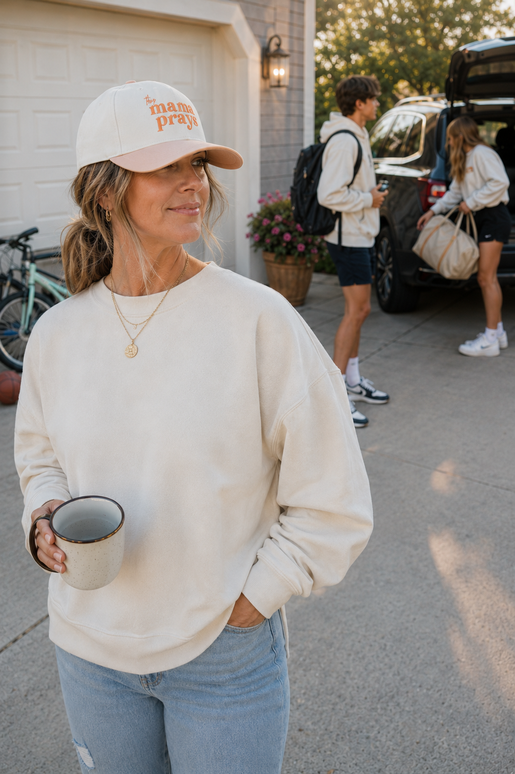 A woman in a cream crewneck sweatshirt and a white baseball cap with orange text stands in a driveway holding a mug. In the background, other people are seen near a car and a bicycle.
