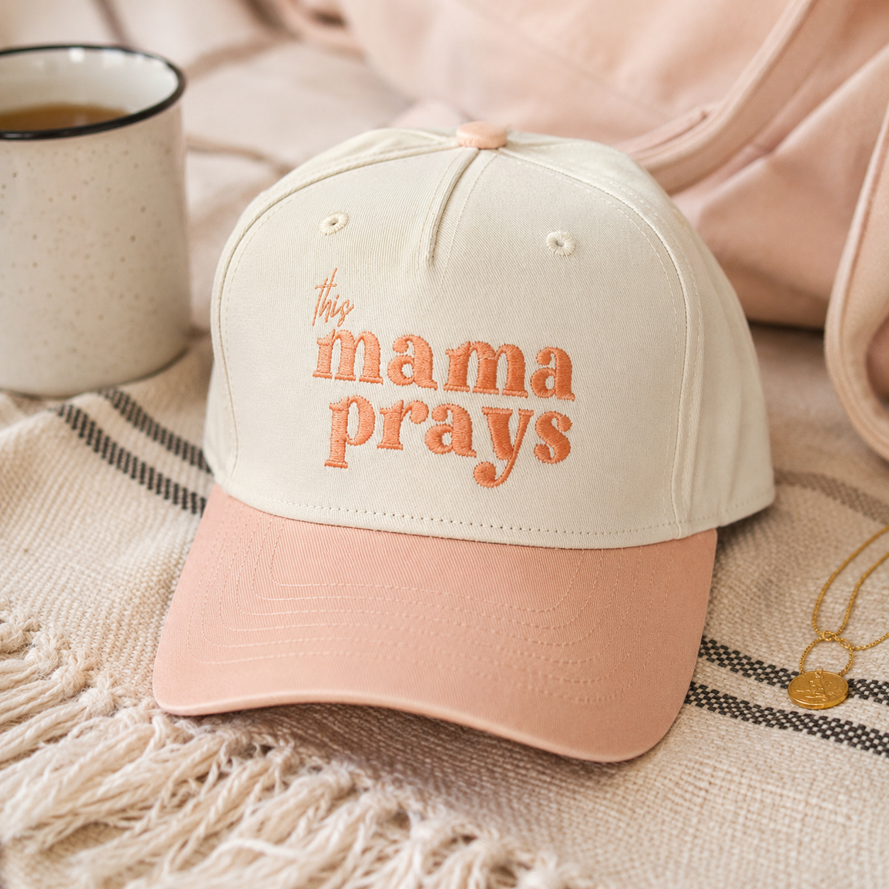 Beige pastel peach cap with text embroidered ‘this mama prays’ set in a cozy blanket with delicate gold jewelry and a cup of coffee around it. 