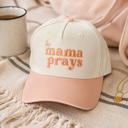 Beige pastel peach cap with text embroidered ‘this mama prays’ set in a cozy blanket with delicate gold jewelry and a cup of coffee around it. 