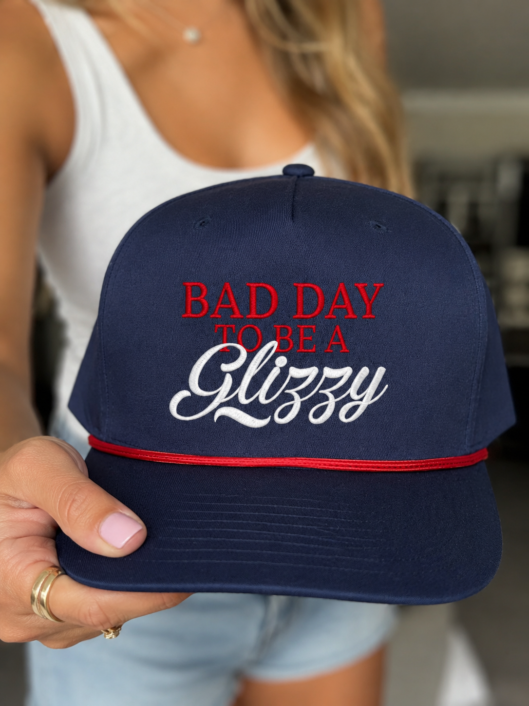 A navy blue baseball cap held by a person, featuring red and white embroidered text that reads 'BAD DAY TO BE A Glizzy' and a red rope across the brim.