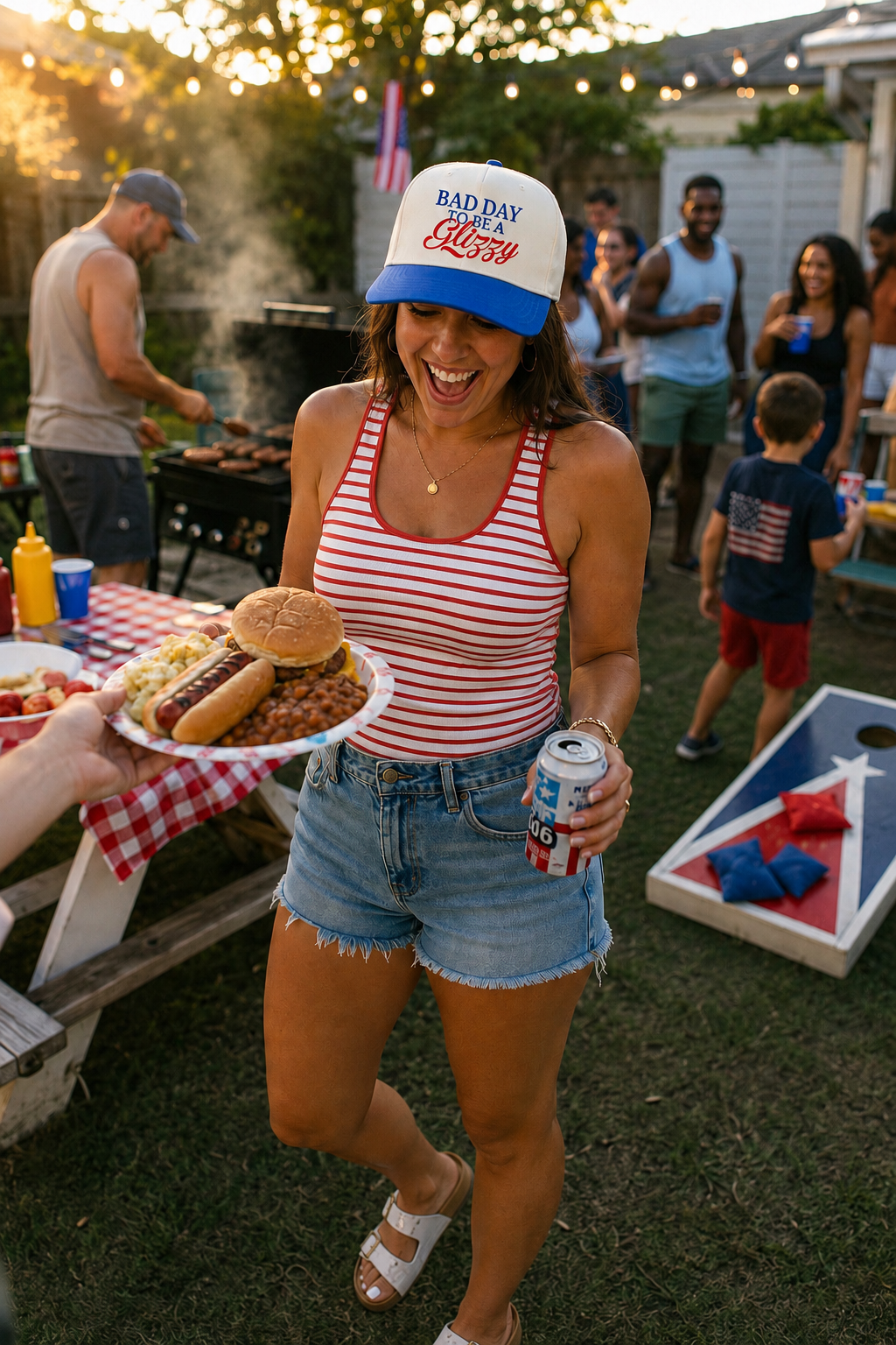 A woman at a backyard BBQ wearing a red and white striped tank top, denim shorts, and a white and blue baseball cap that reads 'BAD DAY FOR A GLIZZY'. She is holding a plate of food and a beverage can, smiling and laughing in the foreground with other people and a cornhole board in the background.
