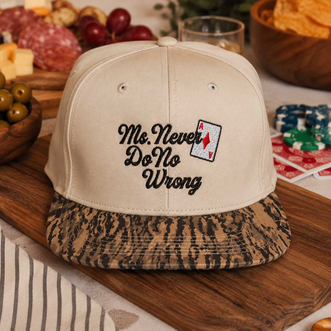 A beige baseball cap with a patterned brown and black visor, featuring black embroidered text that reads 'Ms. Never Do No Wrong' and a small aces card graphic on the front.