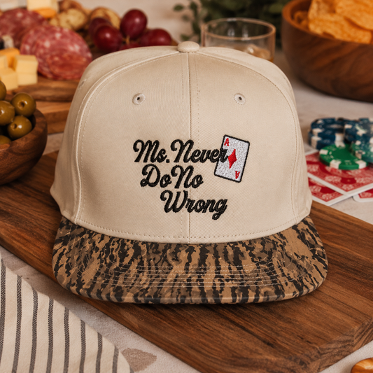 A beige baseball cap with a patterned brown and black visor, featuring black embroidered text that reads 'Ms. Never Do No Wrong' and a small aces card graphic on the front.