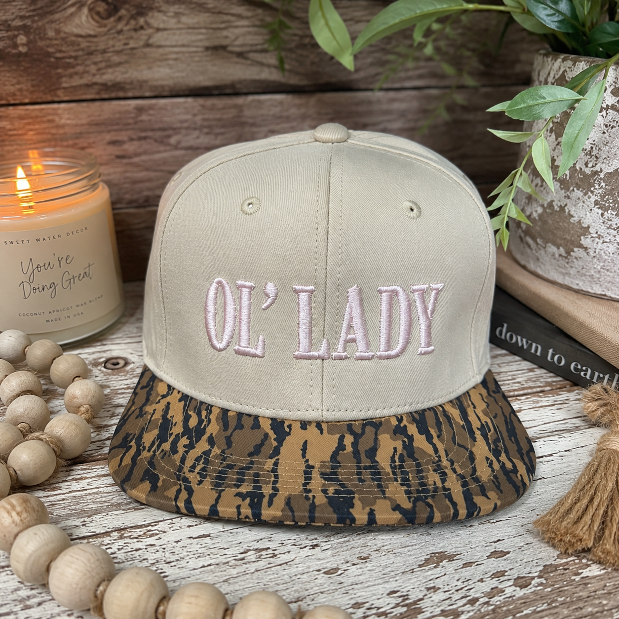 A beige snapback cap with 'OL' LADY' embroidered in pink on the front and a brown/black leopard-print brim, styled on a rustic white wooden surface with a candle, beads, and a plant.