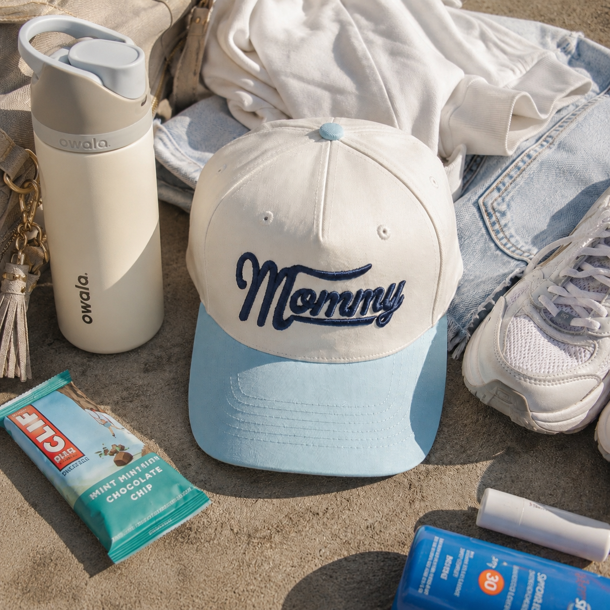 A light blue and natural baseball cap with 'Mommy' embroidered in navy script, styled in a flat lay with a white Owala water bottle, a chocolate chip cookie, a blue sunscreen bottle, and other summer clothing on a concrete surface.
