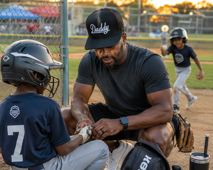 A man wearing a black 'Daddy' cap and dark athletic wear crouches on a baseball field with a young player in a navy jersey, while another child runs in the background.