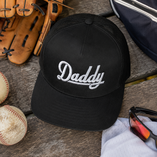 A black trucker-style baseball cap with 'Daddy' embroidered in white script on the front, styled on a wooden surface with baseballs, a glove, sunglasses, and a navy bag around it.