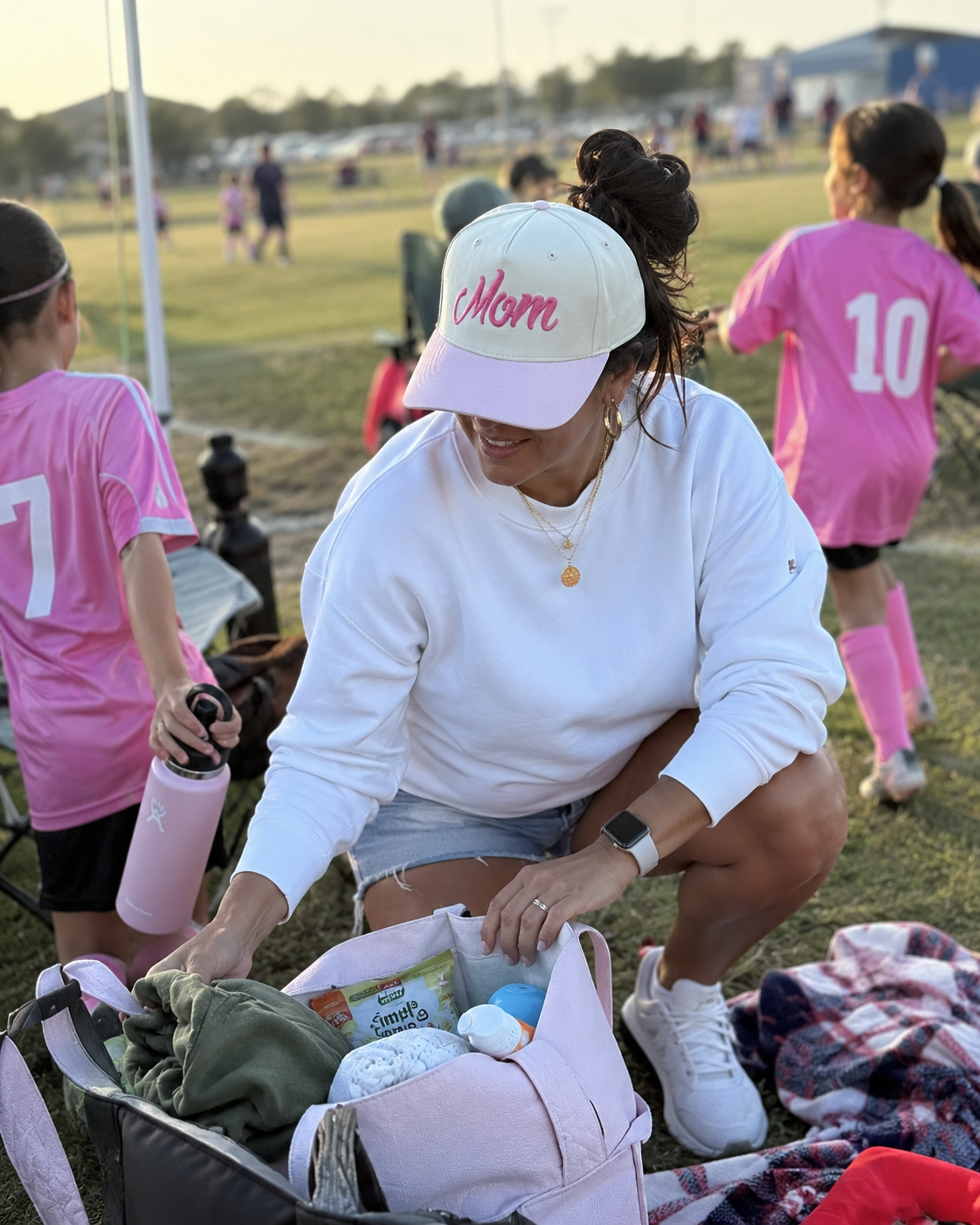 A woman crouches on a soccer field at sunset, organizing a pink diaper bag filled with baby items. She wears a white sweatshirt, denim shorts, white sneakers, and a white cap with pink script reading 'Mom'.