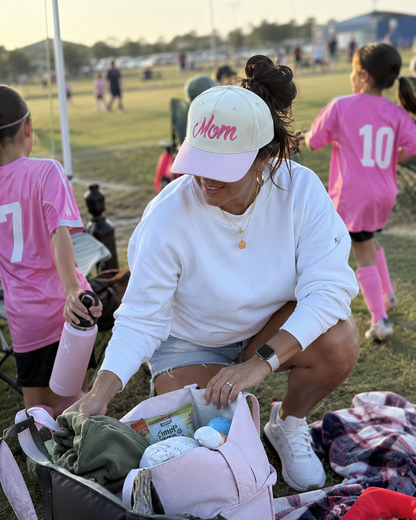 A woman crouches on a soccer field at sunset, organizing a pink diaper bag filled with baby items. She wears a white sweatshirt, denim shorts, white sneakers, and a white cap with pink script reading 'Mom'.