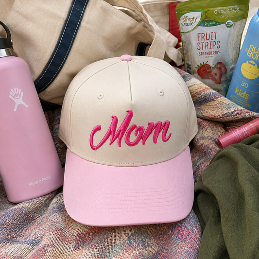 A beige baseball cap with a pink visor and pink embroidered 'Mom' text, styled with a pink Hydro Flask bottle, a beige tote bag, and snack packaging in a cozy flat-lay arrangement.