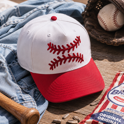 A baseball cap styled on a wooden surface with denim, a baseball in a glove, a bat, sunflower seeds, and a bag of Bunnings. The cap is white on top with red baseball-stitch embroidery, and has a red curved brim.