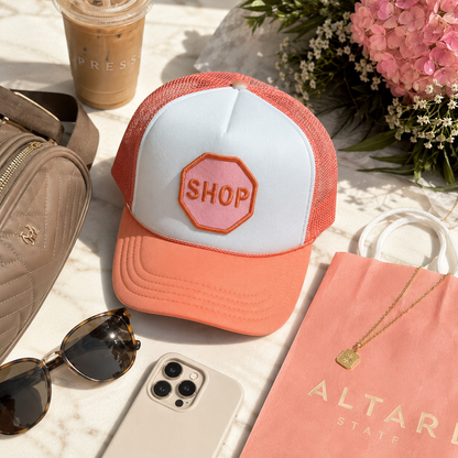 A coral and white trucker hat with a pink 'SHOP' patch on the front, styled in a flat lay with a quilted bag, sunglasses, a phone, flowers, and a pink shopping bag on a marble surface.