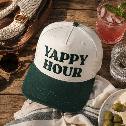 A natural and dark green baseball cap with 'YAPPY HOUR' embroidered on the front, styled on a rustic wooden table with a cocktail, sunglasses, woven sandals, and olives.