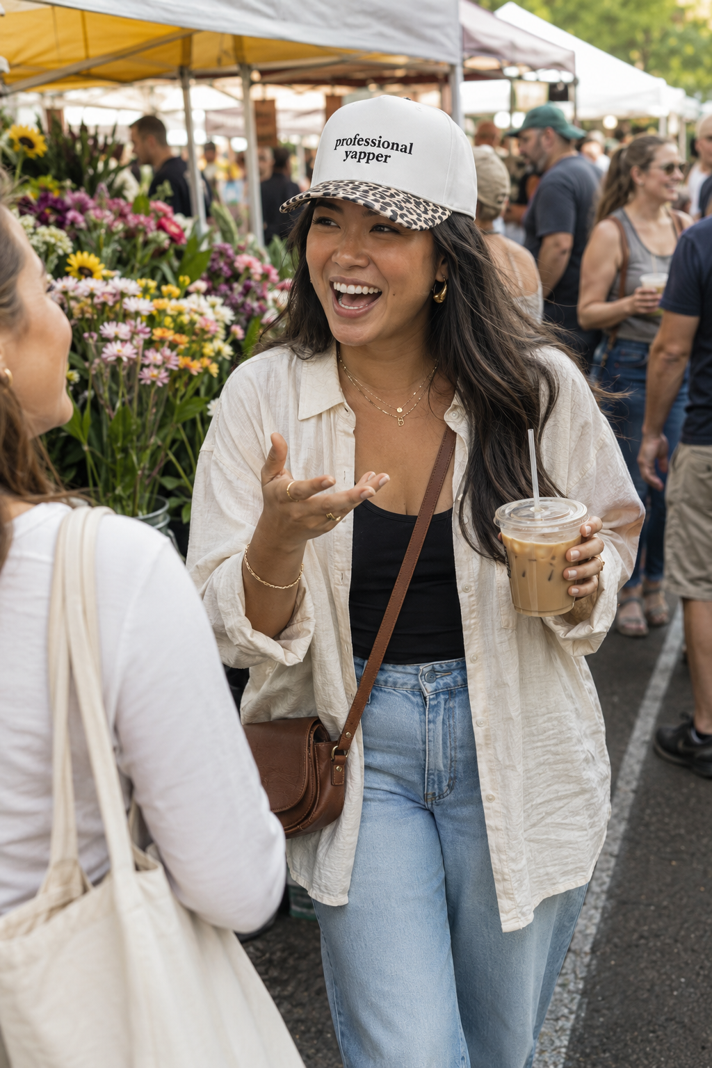 A woman at an outdoor flower market wears a white baseball cap with 'professional shopper' printed on it, a cream button-up shirt, a black tank top, light blue jeans, and carries a brown crossbody bag while holding an iced coffee.