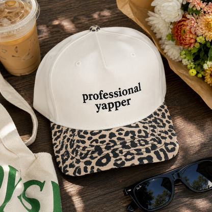 A white baseball cap with black text reading 'professional yapper' and a leopard-print brim, styled on a wooden surface with sunglasses, a tote bag, an iced coffee cup, and flowers nearby.
