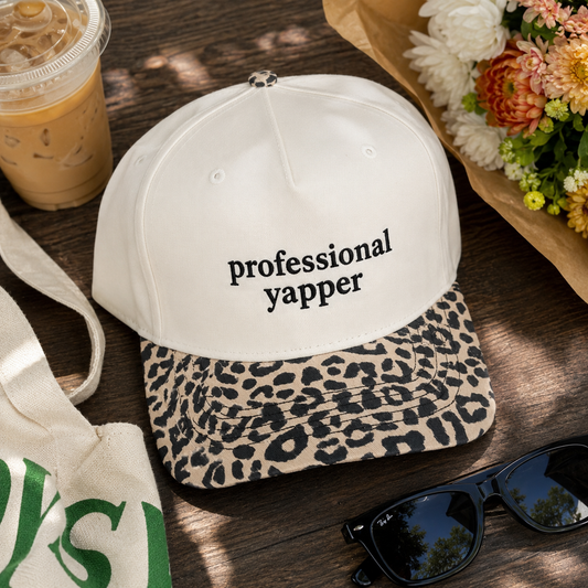 A white baseball cap with black text reading 'professional yapper' and a leopard-print brim, styled on a wooden surface with sunglasses, a tote bag, an iced coffee cup, and flowers nearby.