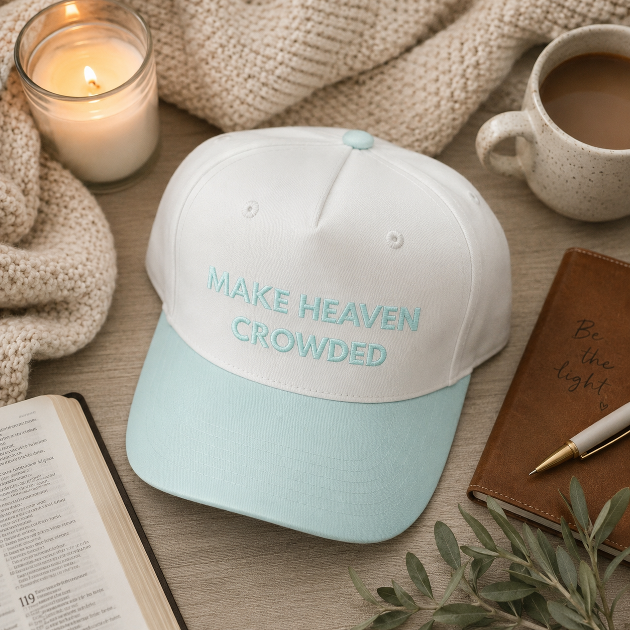 A baseball cap with a off-white crown and a light blue brim, featuring the embroidered text 'MAKE HEAVEN CROWDED' in light blue. The cap is styled in a cozy flat-lay setting with a lit candle, a mug of coffee, a notebook, and a book on a wooden table.