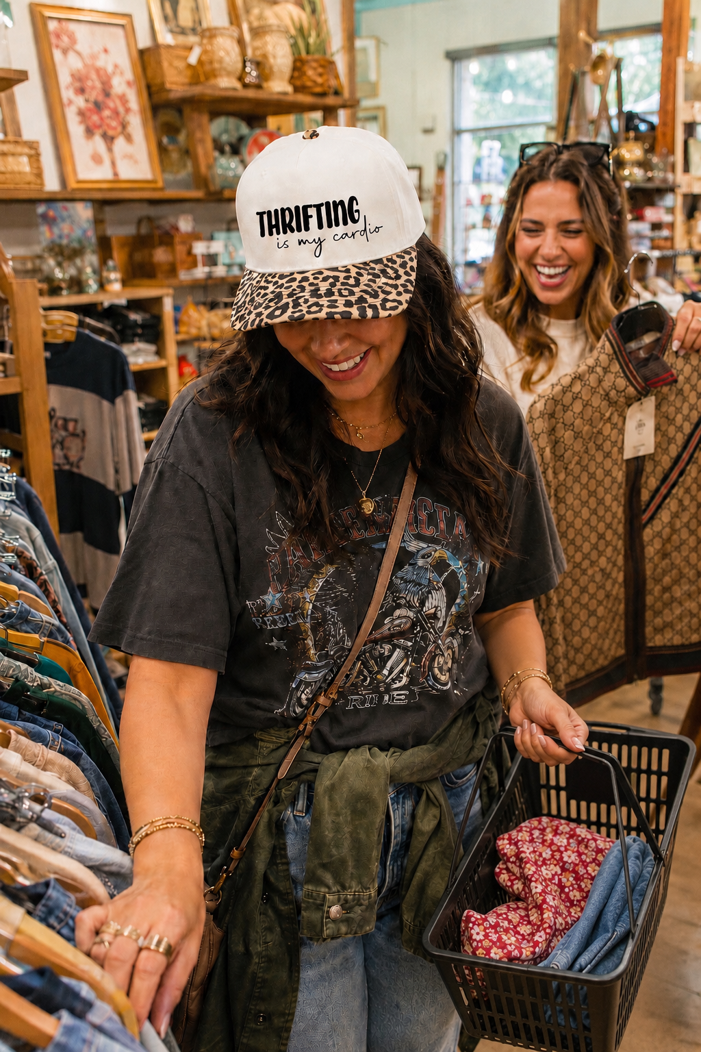 A woman in a thrift store wearing a white 'THRIFTING my craft' baseball cap with a leopard-print brim, looking into clothing racks while holding a black shopping basket. Another woman is visible in the background.