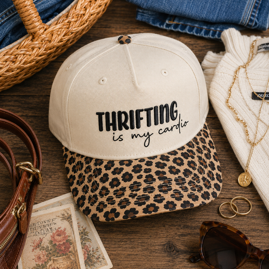 A cream baseball cap with black text reading 'THRIFTING is my cardio' sits on a wooden surface, layered over a leopard-print visor. The cap is styled with a wicker basket, folded jeans, a white knit sweater, gold necklaces, rings, sunglasses, and a brown handbag.
