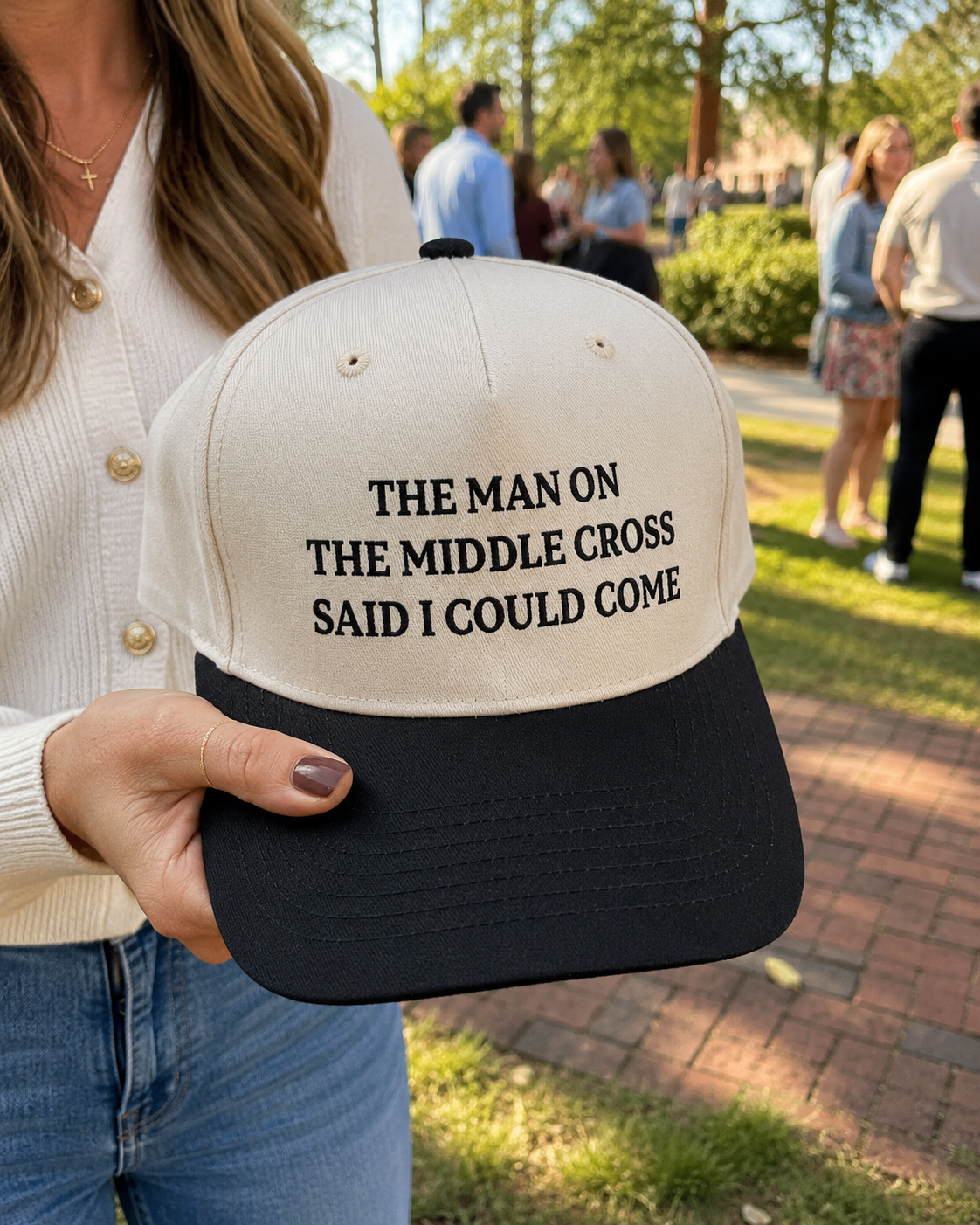A person outdoors holds a black and off-white baseball cap with the text 'THE MAN ON THE MIDDLE CROSS SAID I COULD COME' printed in black on the front. The background shows a blurred park scene with people walking on a brick path.