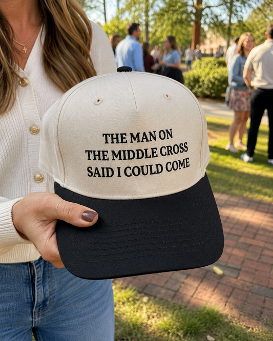 A person outdoors holds a black and off-white baseball cap with the text 'THE MAN ON THE MIDDLE CROSS SAID I COULD COME' printed in black on the front. The background shows a blurred park scene with people walking on a brick path.