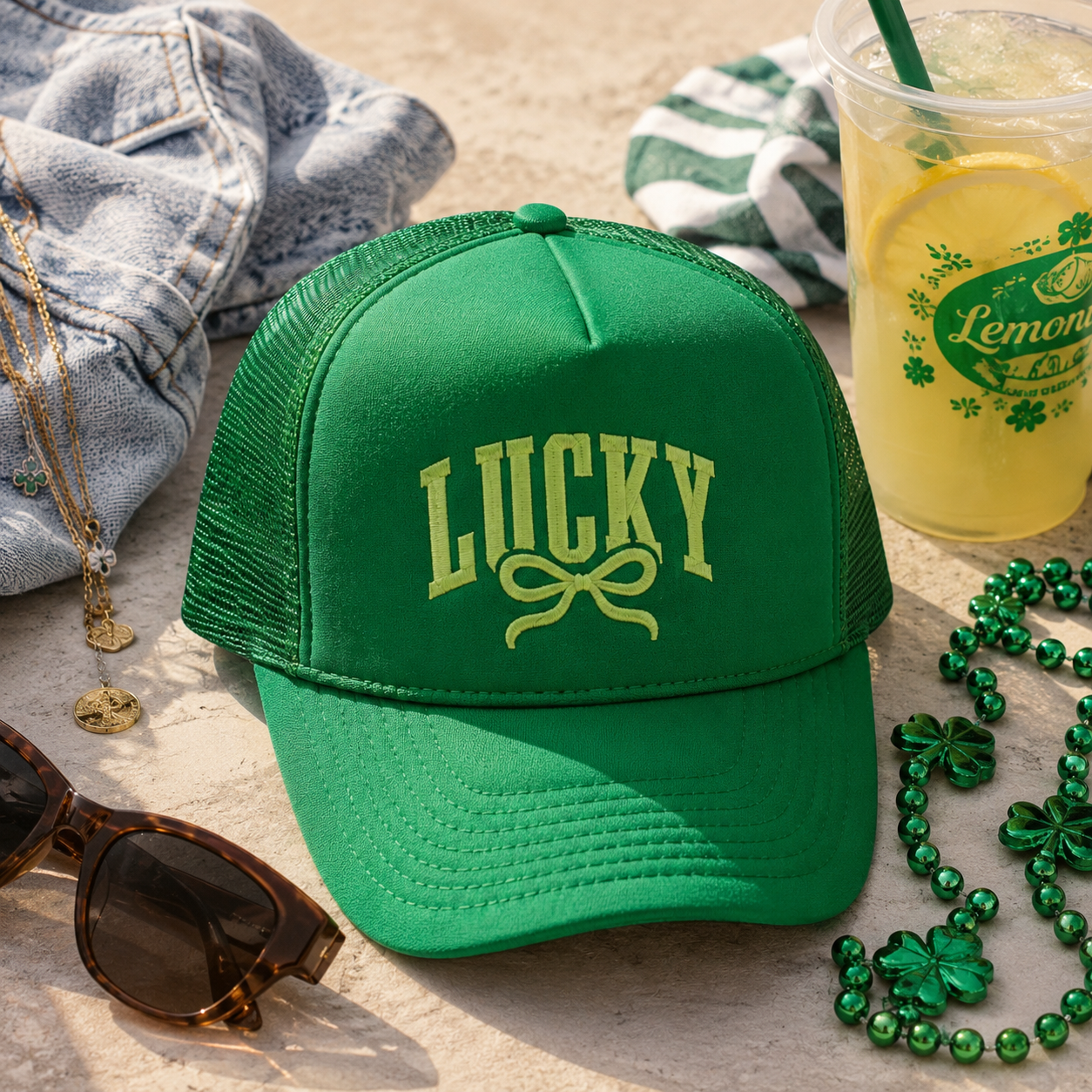 A bright green trucker hat with 'LUCKY' and a bow graphic embroidered on the front, styled on a sandy surface with sunglasses, a denim jacket, a green-and-white striped cloth, a drink, and green shamrock beads.