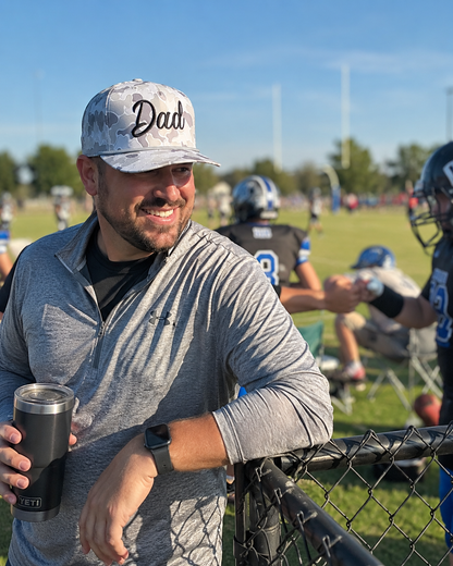 A smiling man in a gray athletic quarter-zip pullover and a gray camouflage baseball cap with 'Dad' written on it, holding a black travel tumbler at an outdoor football practice field.