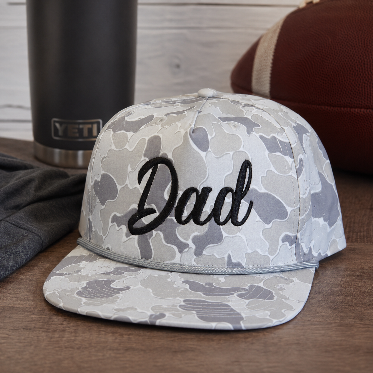 A baseball cap with a gray and white camouflage pattern and the word 'Dad' embroidered in black script on the front, displayed on a wooden surface with a football, a dark sweatshirt, and a black YETI tumbler in the background.